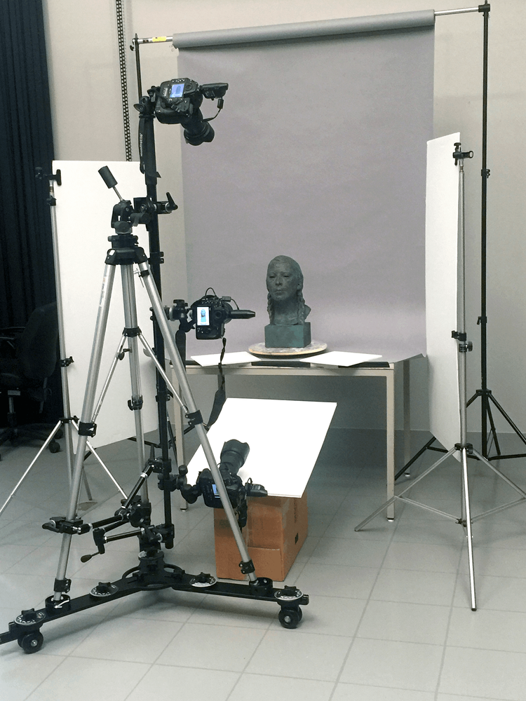Le buste d'une jeune femme est installé sur une table et entouré d'équipement pour une séance de photographie (caméras, trépieds, réflecteurs).