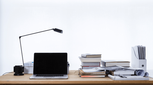 Table de travail en bois sur laquelle se trouve une lampe sur pied, un ordinateur portable de même qu'une une pile de livres et de cahiers.