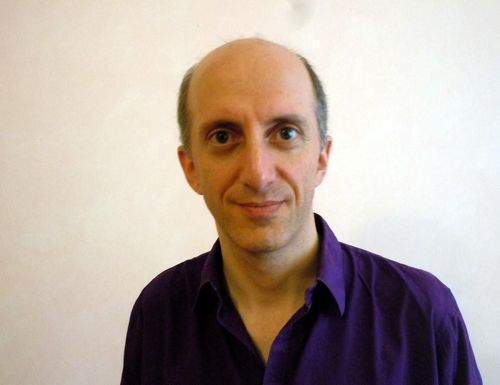 Portrait d'un homme souriant n'ayant pas de cheveux sur le haut du crâne et portant une chemise de couleur violet.  
